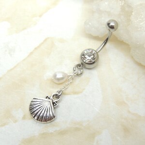 May include: A silver belly button ring with a curved barbell design. The top ball and center gem are clear. A small pearl and a seashell charm dangle from the center gem, connected by a delicate chain. The seashell charm is detailed with ribbed lines.