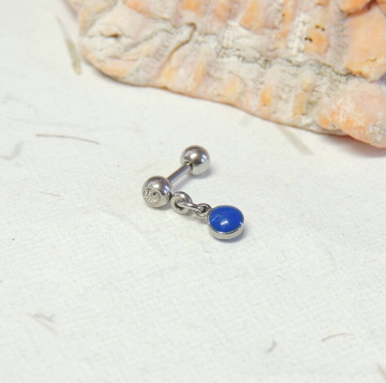 May include: A silver barbell earring featuring a small, round, blue gemstone pendant. The earring has a ball at each end of the post, with a small, clear stone on one end. The blue charm is a vibrant, solid colour.