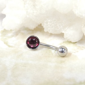 May include: A curved barbell body piercing with a round, deep red gemstone on one end and a small silver ball on the other. The jewelry is made of silver-colored metal.