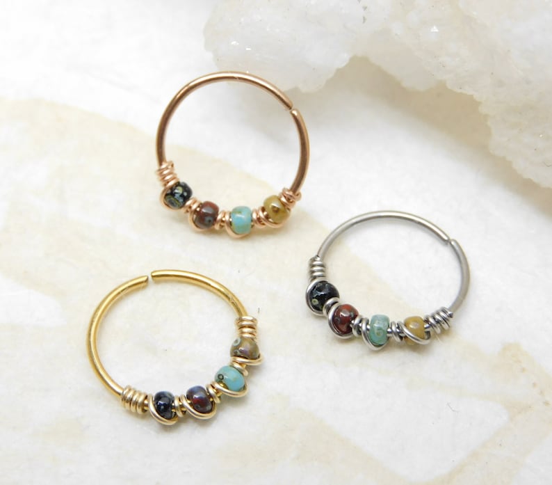 May include: Three nose rings with colourful beads. The rings are made of silver, gold, and rose gold. The beads are a mix of black, brown, turquoise, and gold.
