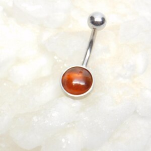 Puede incluir: Un piercing de ombligo plateado con una gema redonda de color naranja.