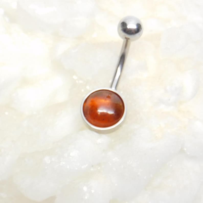 Puede incluir: Un piercing de ombligo plateado con una gema redonda de color naranja.