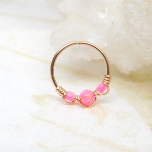 May include: A rose gold nose ring featuring three pink opal beads. The beads are wire-wrapped and set on a circular hoop. The jewelry is designed for nose piercings and is a delicate accessory.