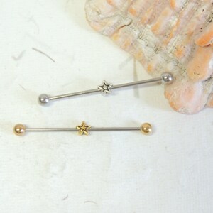 May include: Two barbell style body piercings with star-shaped charms. The top barbell is silver with a silver star charm. The bottom barbell is gold with a gold star charm.