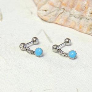 May include: A pair of silver barbell earrings, each with a small, light blue bead suspended by a chain. One earring features a small, clear gem on the barbell end. The earrings are presented on a textured, off-white surface.
