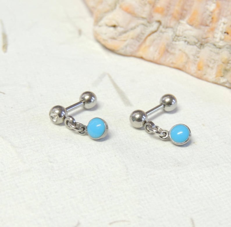 May include: A pair of silver barbell earrings, each with a small, light blue bead suspended by a chain. One earring features a small, clear gem on the barbell end. The earrings are presented on a textured, off-white surface.