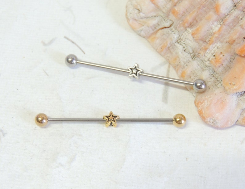 May include: Two silver barbell style body piercings with star-shaped charms. The top barbell has a silver charm and the bottom barbell has a gold charm.