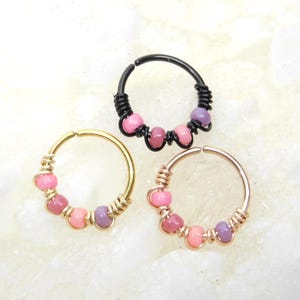 May include: Three beaded hoop earrings in gold, black, and rose gold. Each earring features a series of pink and purple beads. The hoops are wrapped with wire and have a simple, elegant design. The earrings are displayed on a white surface.
