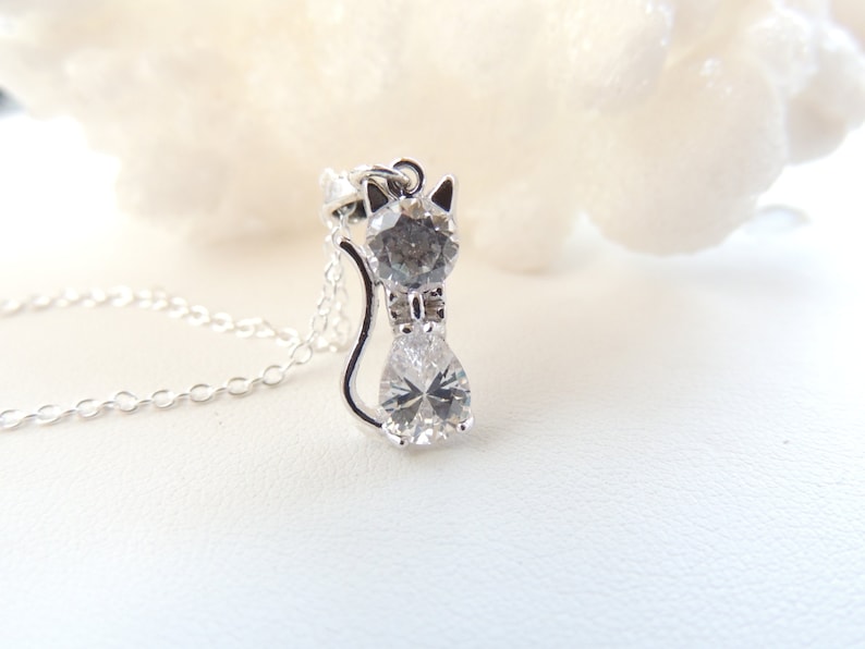 Puede incluir: Un collar con colgante de gato plateado con un cuerpo de cristal transparente en forma de pera y una cabeza de cristal gris. Las orejas del gato son puntiagudas y la cadena es delicada. El colgante est&aacute; sobre una superficie blanca.