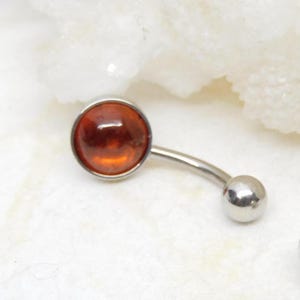 Puede incluir: Un piercing de ombligo plateado con una gema redonda de &aacute;mbar. El anillo es curvo y tiene una peque&ntilde;a bola plateada en el extremo.
