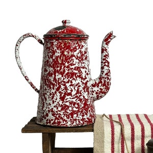 Può includere: Una caffettiera vintage in smalto rosso e bianco maculato è appoggiata su uno sgabello di legno. Un runner in lino a righe rosse e bianche e un gomitolo di spago sono accanto a una piccola caffettiera in metallo. Arredamento rustico della cucina.