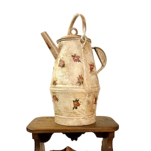 May include: A cream-coloured metal teapot with a handle and lid, decorated with hand-painted red and pink roses. It rests on a small, weathered wooden stool, alongside a pair of wooden clogs and antique books. The image presents a rustic, antique aesthetic.