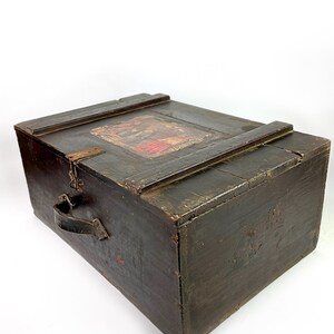May include: A dark wooden chest with a hinged lid and a leather handle. The lid has a faded, colorful label with an image of people.