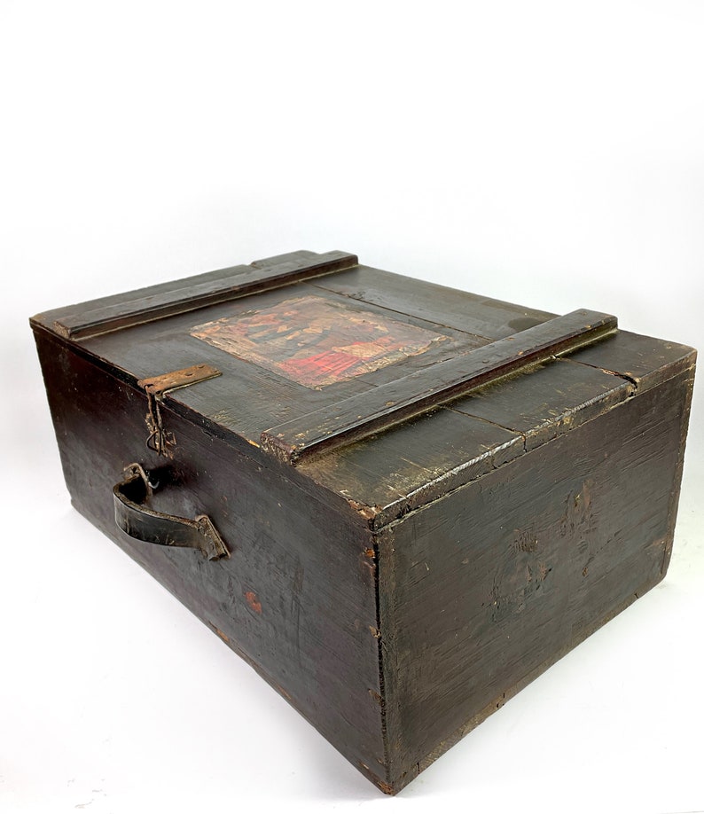 May include: A dark wooden chest with a hinged lid and a leather handle. The lid has a faded, colorful label with an image of people.