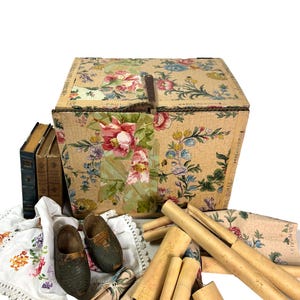 May include: A vintage floral-patterned box with a metal clasp, surrounded by antique books, wooden clogs, rolled papers, and a floral embroidered cloth. The box features a beige background with pink, blue, and green floral designs.