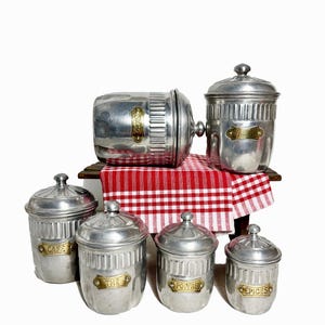 Puede incluir: Un conjunto de botes metálicos vintage con tapas, etiquetados como "CAFE", "THE", "POIVRE" y "EPICES". Los botes están colocados sobre una pequeña mesa de madera cubierta con un paño a cuadros rojos y blancos. Los botes tienen un acabado plateado.