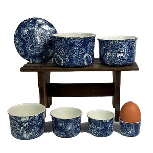 Peut inclure: Un ensemble de bols en céramique bleu et blanc et un coquetier avec un motif floral. Les bols sont de tailles différentes et sont disposés sur une étagère en bois. Un œuf est dans le coquetier.