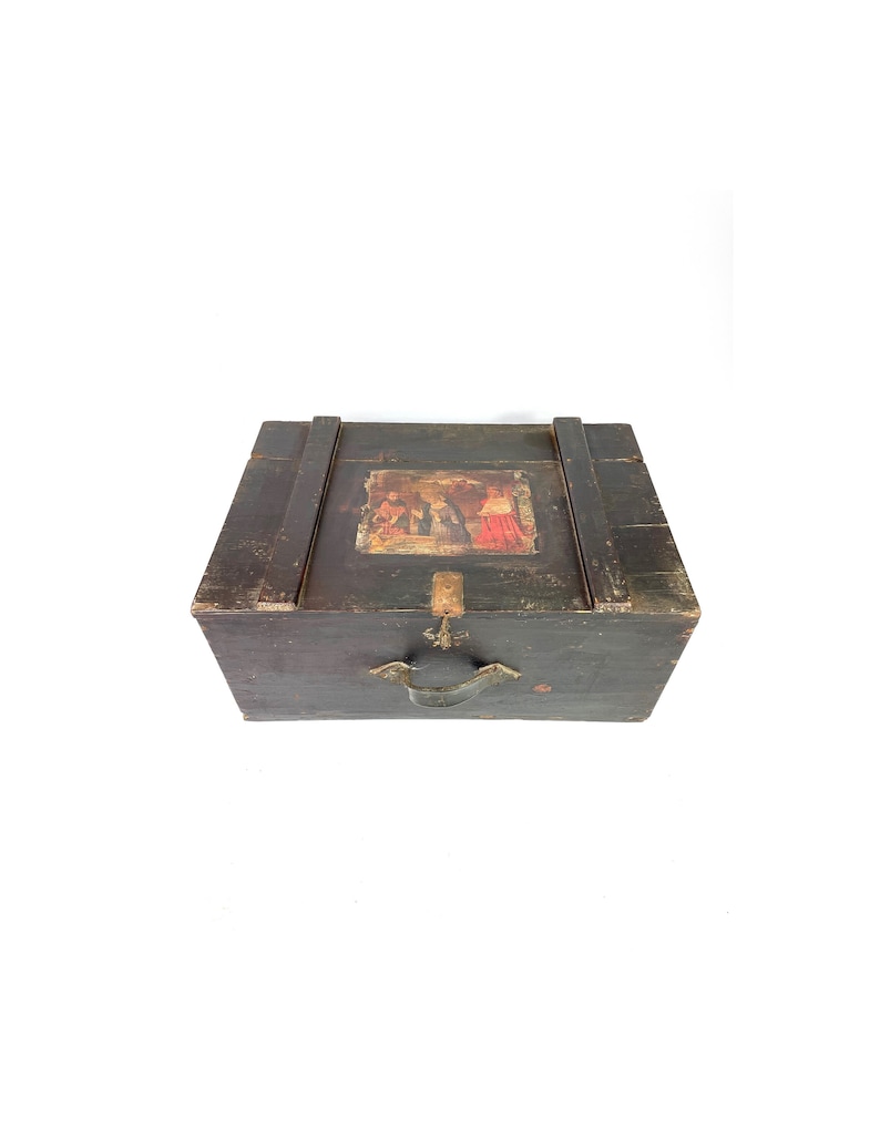 May include: A dark wooden chest with a hinged lid and a metal handle. The chest has a painted scene on the top with figures in red and gold.