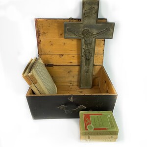 May include: A wooden box with a black lid and a metal cross with a figure of Jesus Christ on it. The box is open and contains a stack of books. The books are wrapped in brown paper and have a green cover with gold lettering. The cross is made of metal and has a dark patina. The box is sitting on a white surface.