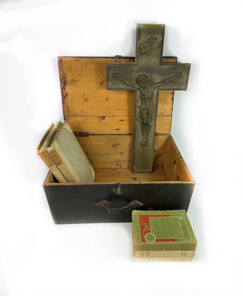 May include: A wooden box with a black lid and a metal cross with a figure of Jesus Christ on it. The box is open and contains a stack of books. The books are wrapped in brown paper and have a green cover with gold lettering. The cross is made of metal and has a dark patina. The box is sitting on a white surface.