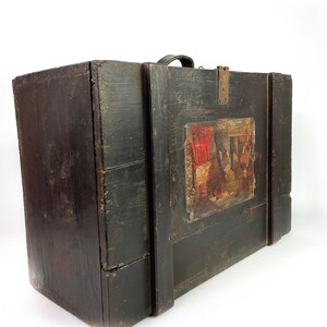 May include: A vintage wooden box with a black painted finish and a leather handle. The box has a painted scene on the front with red, yellow, and blue colors.