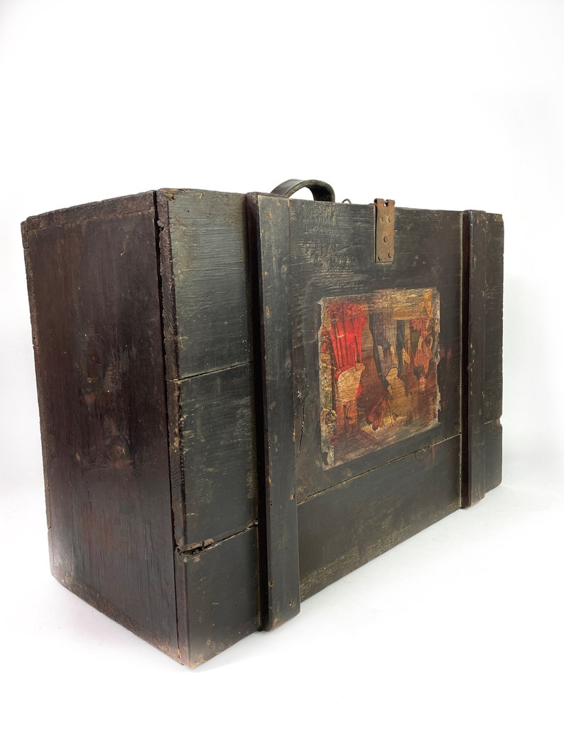 May include: A vintage wooden box with a black painted finish and a leather handle. The box has a painted scene on the front with red, yellow, and blue colors.