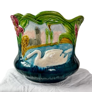 May include: A ceramic planter with a swan design. The planter features a scalloped rim with green foliage and pink flowers. The body of the planter is a deep teal color, with a painted scene of a swan on a lake.