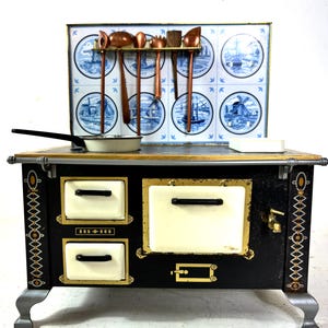Könnte beinhalten: Ein schwarzer und goldener Spielzeugherd im Vintage-Stil mit cremefarbenen Akzenten und einer Delft-Fliesen-Rückwand. Der Herd verfügt über eine kleine Pfanne, ein weißes Geschirr und Kochutensilien aus Kupfer. Die Rückwand zeigt blau-weiße Bilder von Windmühlen und Booten.