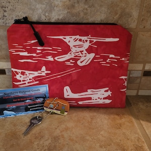 Puede incluir: Bolsa roja con cremallera con un estampado blanco de hidroaviones volando sobre el agua. La bolsa está junto a una tarjeta de visita que dice "Northern Outpost Goods" y un llavero con un pequeño amuleto de casa.