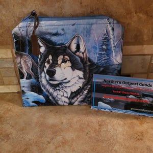 May include: A blue zippered pouch with a wolf and trees design. The pouch is made by Northern Outpost Goods. The text on the business card reads "Inspired by Handmade Goods. Visit My Outpost Shop At:  northernoutpostgoods.com"