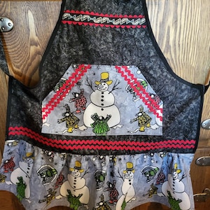 May include: A black and gray apron with a winter theme. The apron features a large pocket with a snowman design and red trim. The bottom of the apron has a blue and white pattern with snowmen and a red ruffle.