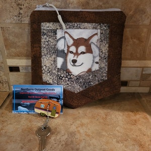 May include: A brown fabric pouch with a zipper closure and a pocket featuring a white and brown husky dog face. The pouch is on a tiled surface with a business card and a key chain with a camper van charm.