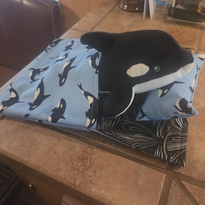 May include: A plush orca toy resting on a small pillow and bed set. The bedding features a light blue fabric with a repeating pattern of black and white orcas, while the base of the bed is made of a black fabric with a white swirling line pattern.