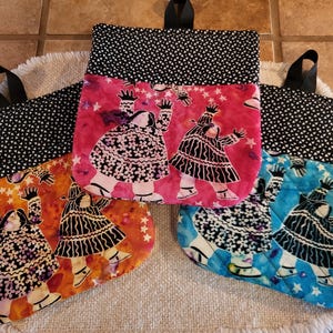 Op de afbeelding: Vier gewatteerde ovenwanten met zwarte ophanglussen. De wanten hebben een zwart-wit sterrenpatroon aan de bovenkant en een grillig ontwerp van figuren in kleding met patronen op een levendige achtergrond van oranje, roze en blauw.