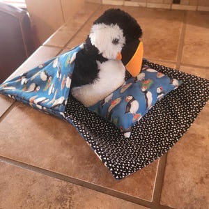 May include: A plush puffin toy resting on a small rectangular pillow, tucked into a fabric sleeping bag. The sleeping bag and pillow feature a blue fabric with a puffin pattern, while the base fabric has a black and white geometric print.