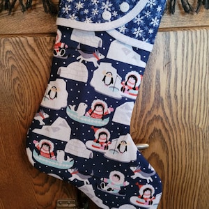 May include: A blue Christmas stocking with a white snowflake pattern and a white cuff. The stocking features a variety of winter-themed illustrations, including penguins, igloos, and people wearing winter clothing.