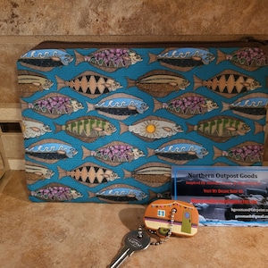 May include: A blue zippered pouch with a repeating pattern of stylised fish. The pouch is made of fabric and has a brown zipper. The pouch is sitting on a tiled surface. A business card for Northern Outpost Goods is in the foreground. The business card has the text "Northern Outpost Goods" and "Inspired by Northern Rivers & Sea Air".