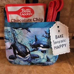 Può includere: Un organizer in tessuto con un motivo di orche blu e bianche, contenente un pacchetto di mix Betty Crocker Chocolate Chip e cucchiai dosatori rossi. Un'etichetta recita "Bake Someone HAPPY".