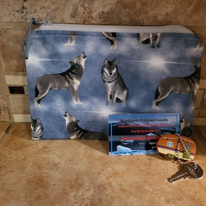 May include: A blue zippered pouch with a wolf print design. The pouch is sitting on a brown tiled surface next to a business card that reads "Northern Outpost Goods" and a key chain with a small camper trailer charm.