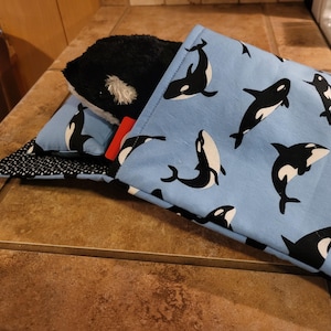 May include: A plush black and white orca stuffed animal peeks out of a light blue fabric pouch. The pouch is patterned with black and white orca illustrations. A smaller pillow with the same orca pattern is visible.