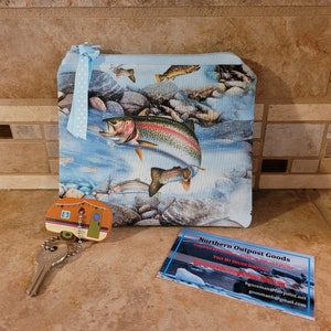 May include: A blue zippered pouch with a colorful image of trout swimming in a stream. The pouch is decorated with a blue ribbon. The pouch is next to a small camper keychain and a business card for Northern Outpost Goods.