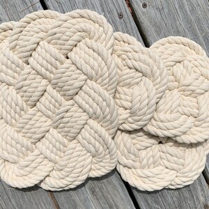 May include: Two off-white, woven rope mats with a braided design. The mats are made of thick, natural-colored rope and are arranged on a weathered wooden surface. The mats have a circular shape with a square center.