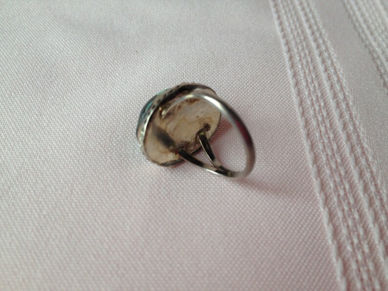 May include: A silver ring with a turquoise stone set in the center. The ring is on a white fabric background.