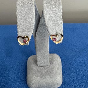 May include: A pair of gold-toned heart-shaped stud earrings. Each earring features a small, round, red gemstone set in the center of the heart. The earrings are displayed on a gray suede jewelry stand.