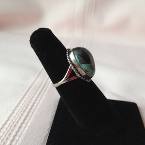 May include: A silver ring with a turquoise stone set in a bezel setting. The ring is on a black ring display stand.