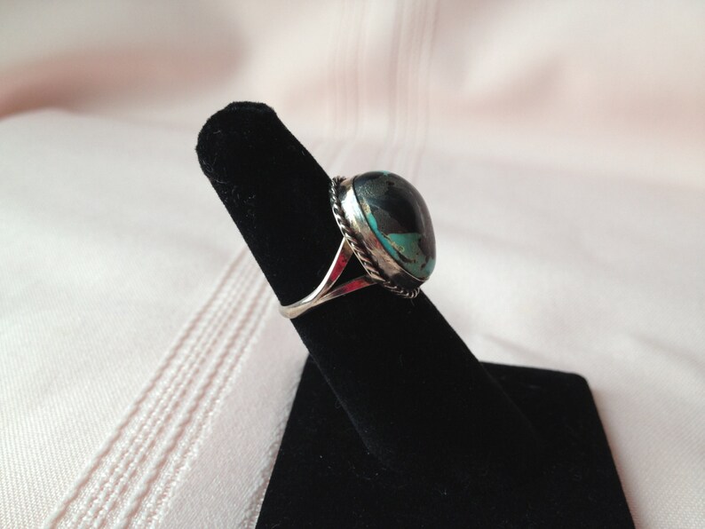 May include: A silver ring with a turquoise stone set in a bezel setting. The ring is on a black ring display stand.