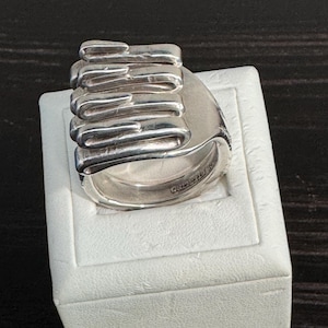 May include: A sterling silver ring featuring a sculptural design of five stacked, horizontal, ribbon-like loops. The metal has a polished, reflective finish with visible indentations and the word STERLING stamped on the inner band surface.