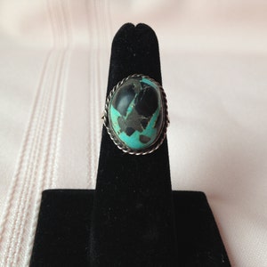 May include: A silver ring with a turquoise and black stone set in a rope-like border.