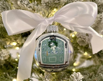 May include: A clear glass Christmas bauble with a painted image of a green door with a wreath and a white bow. The bauble is decorated with a white satin ribbon and the text "Fondamente" is written on the bottom.