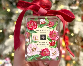 May include: A rectangular cologne bottle with a red cap and ribbon. The bottle is decorated with pink and red floral designs and the text "JO MALONE LONDON". The label reads "PEONY & BLUSH SUEDE COLOGNE".